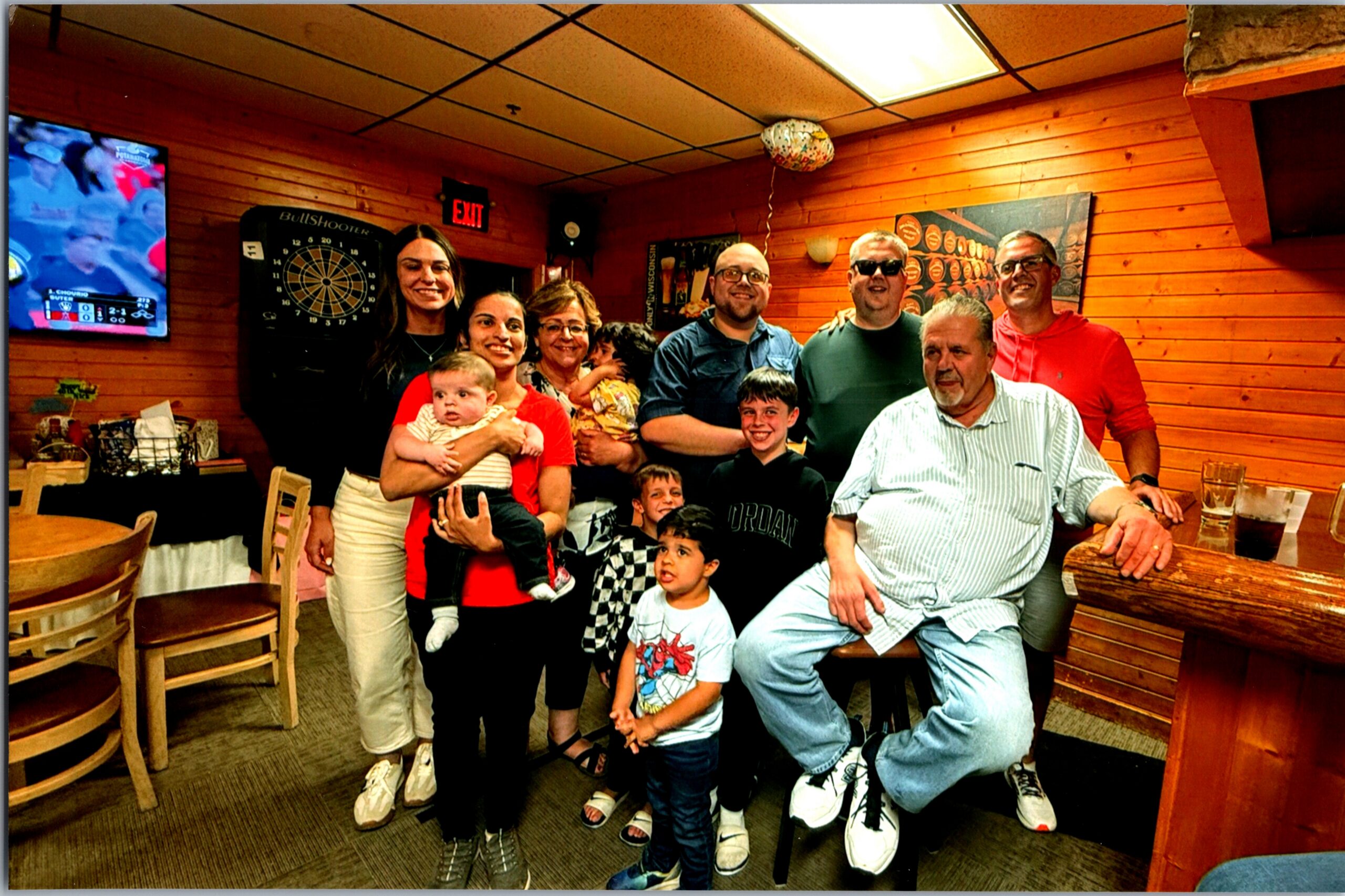 Group of adults and children posing together in a wood-paneled bar/restaurant, with a dartboard on the wall and a balloon overhead.