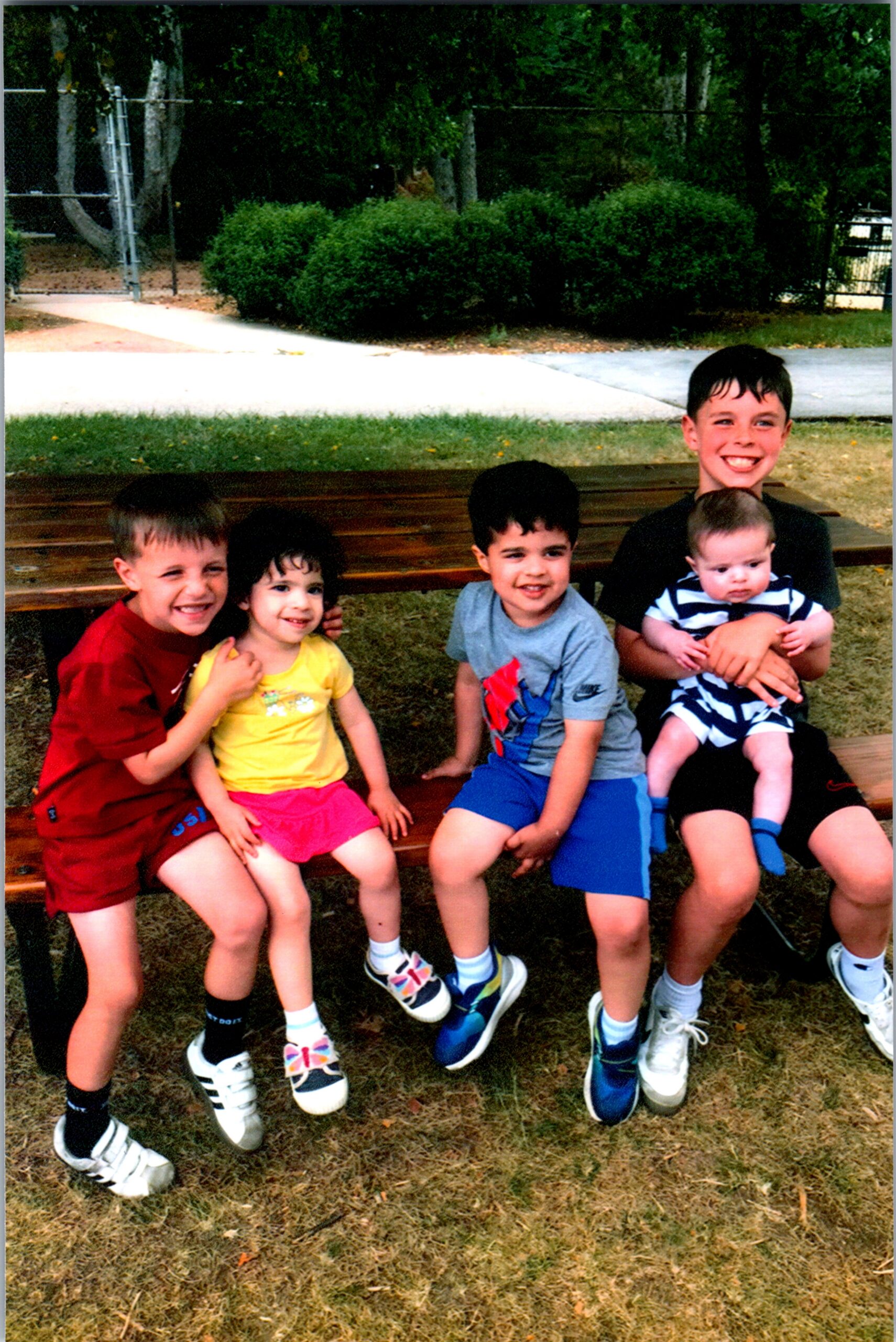 Five children sit close together on a wooden bench outdoors, smiling at the camera on a sunny day.