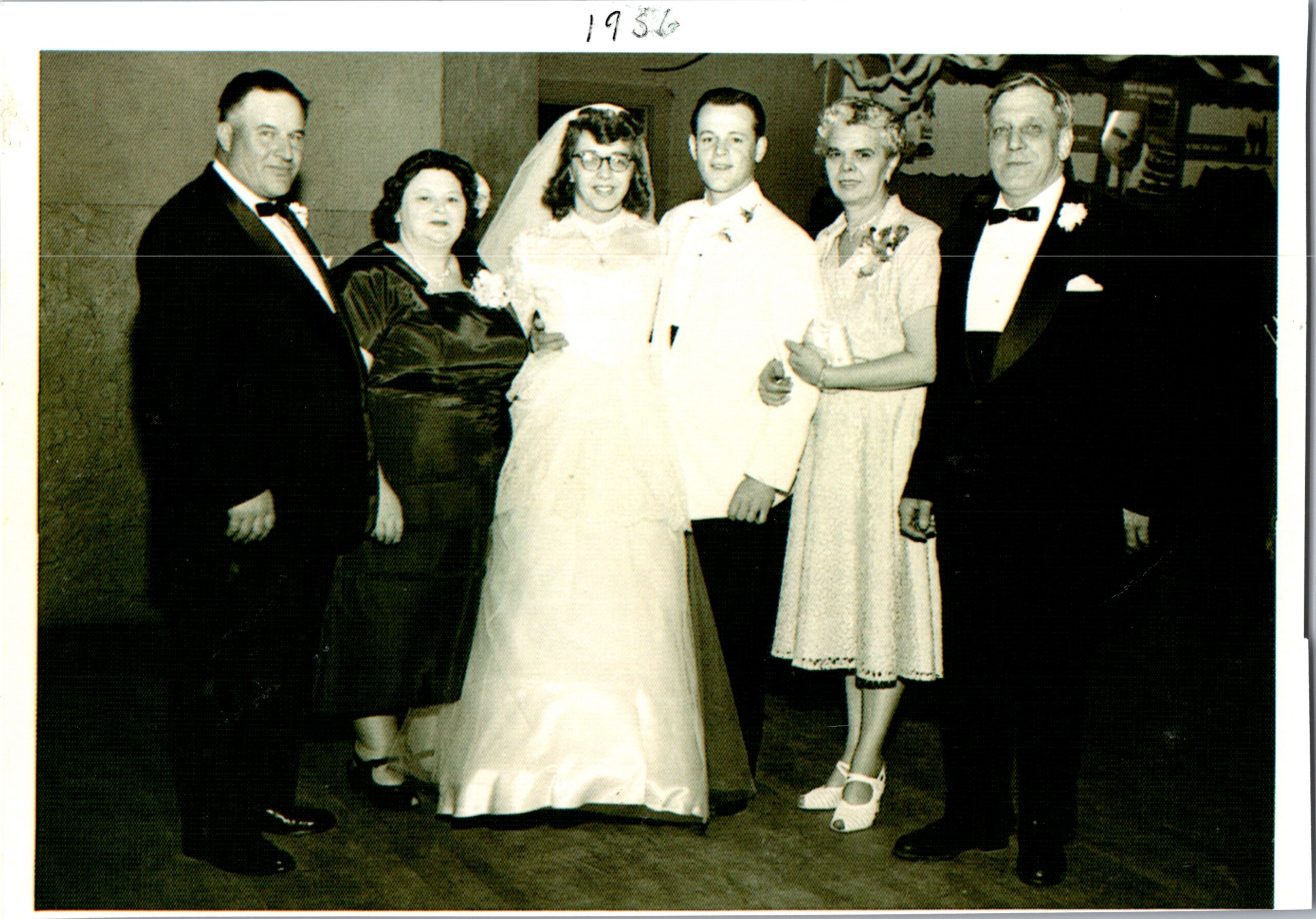 Group of six people at a wedding: bride in white gown and veil, groom in light suit, two bridesmaids in light dresses, and two groomsmen in formal tuxedos posing together.