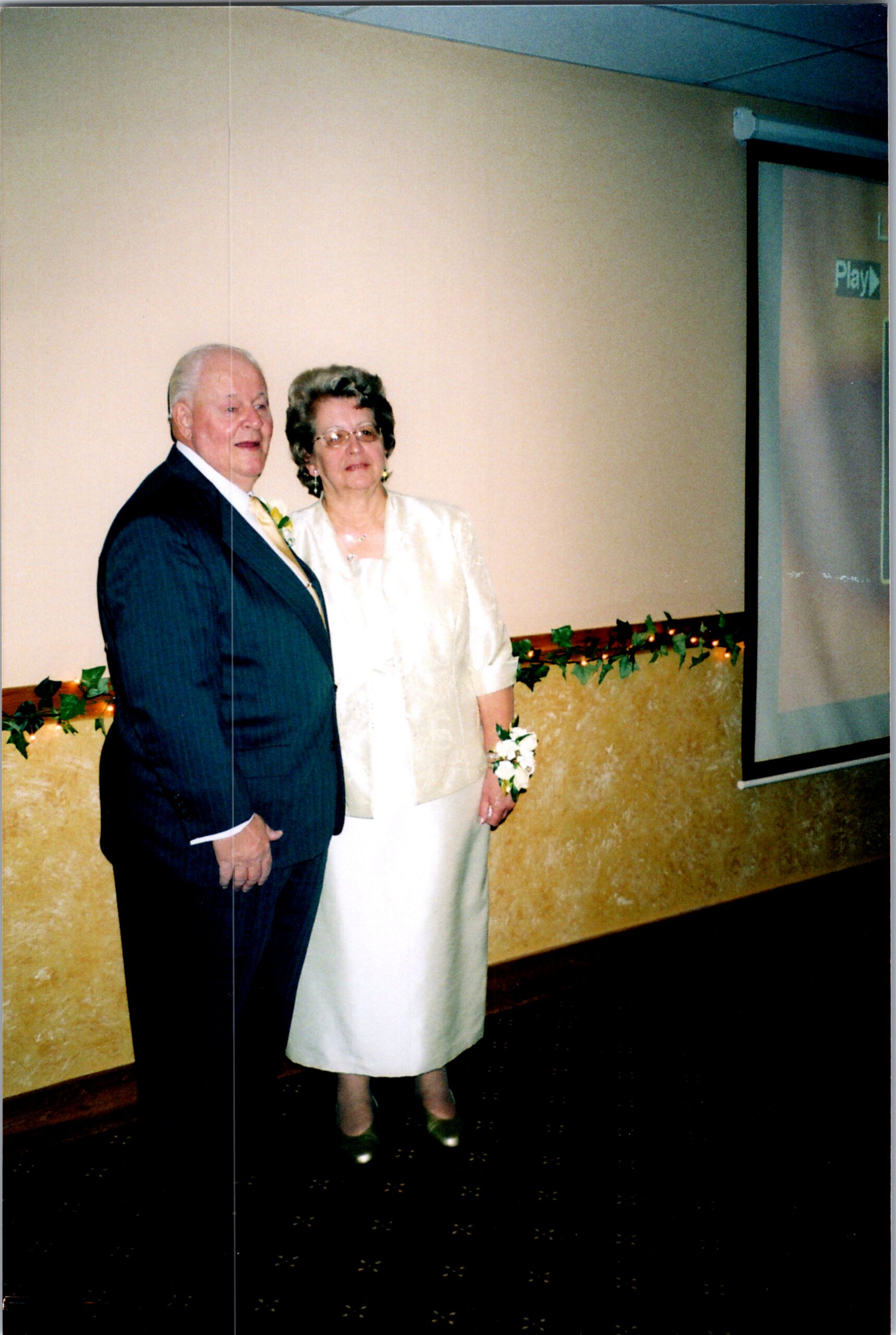 Older couple in formal attire posing together at an indoor event, with greenery along the wall behind them.