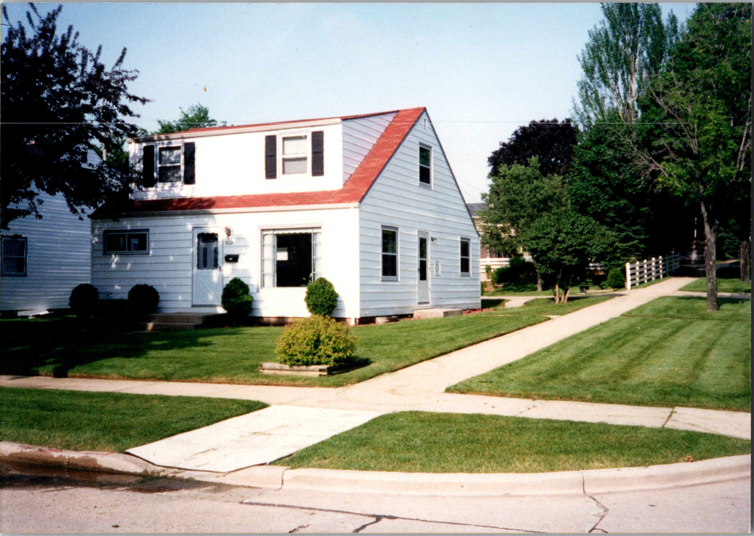 White two-story house with a red pitched roof, black shutters, and a manicured lawn along a sunny street.
