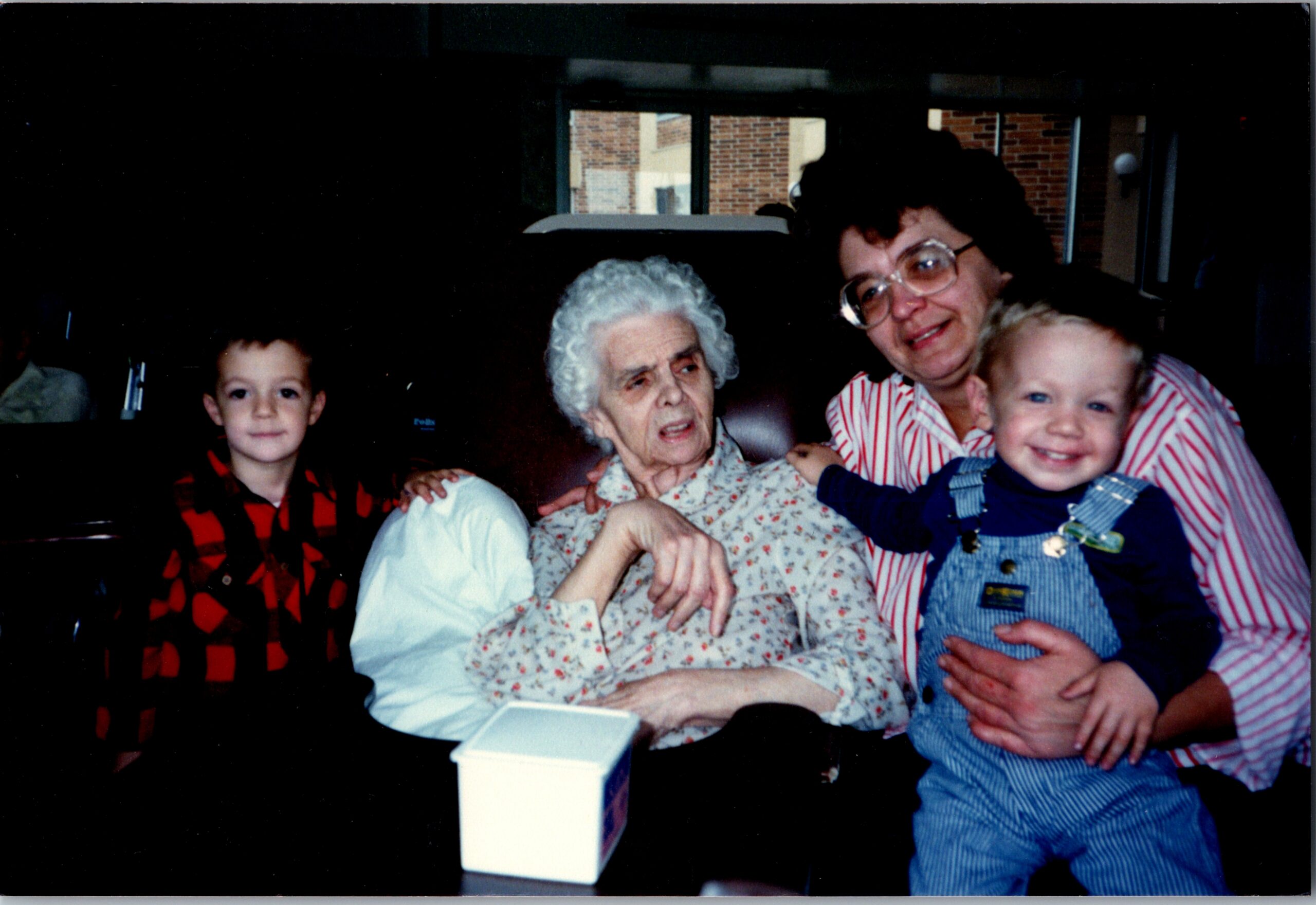 Older woman with white hair and floral blouse sits between a smiling woman in striped shirt and two young children, posing together indoors with a white box on a table in front.