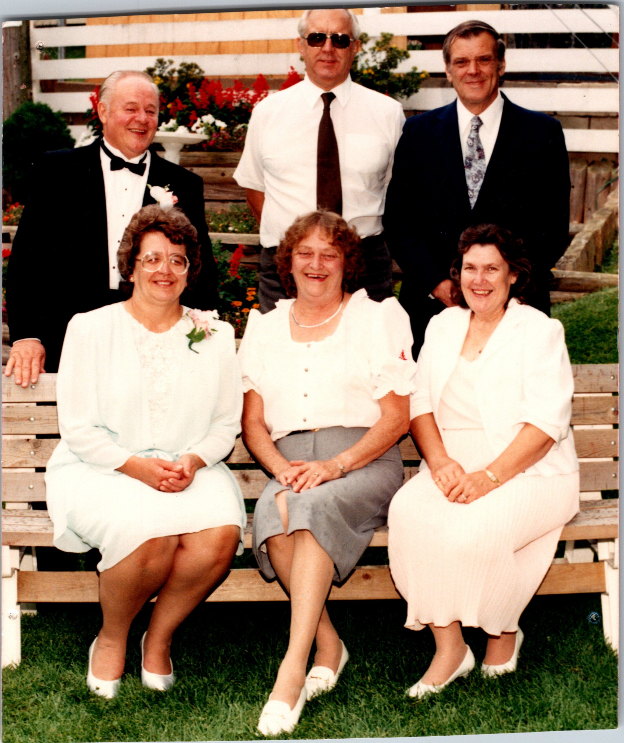 Five older adults posing on a wooden bench outdoors in a garden: three men in formal attire standing behind, two women in light dresses seated in front.