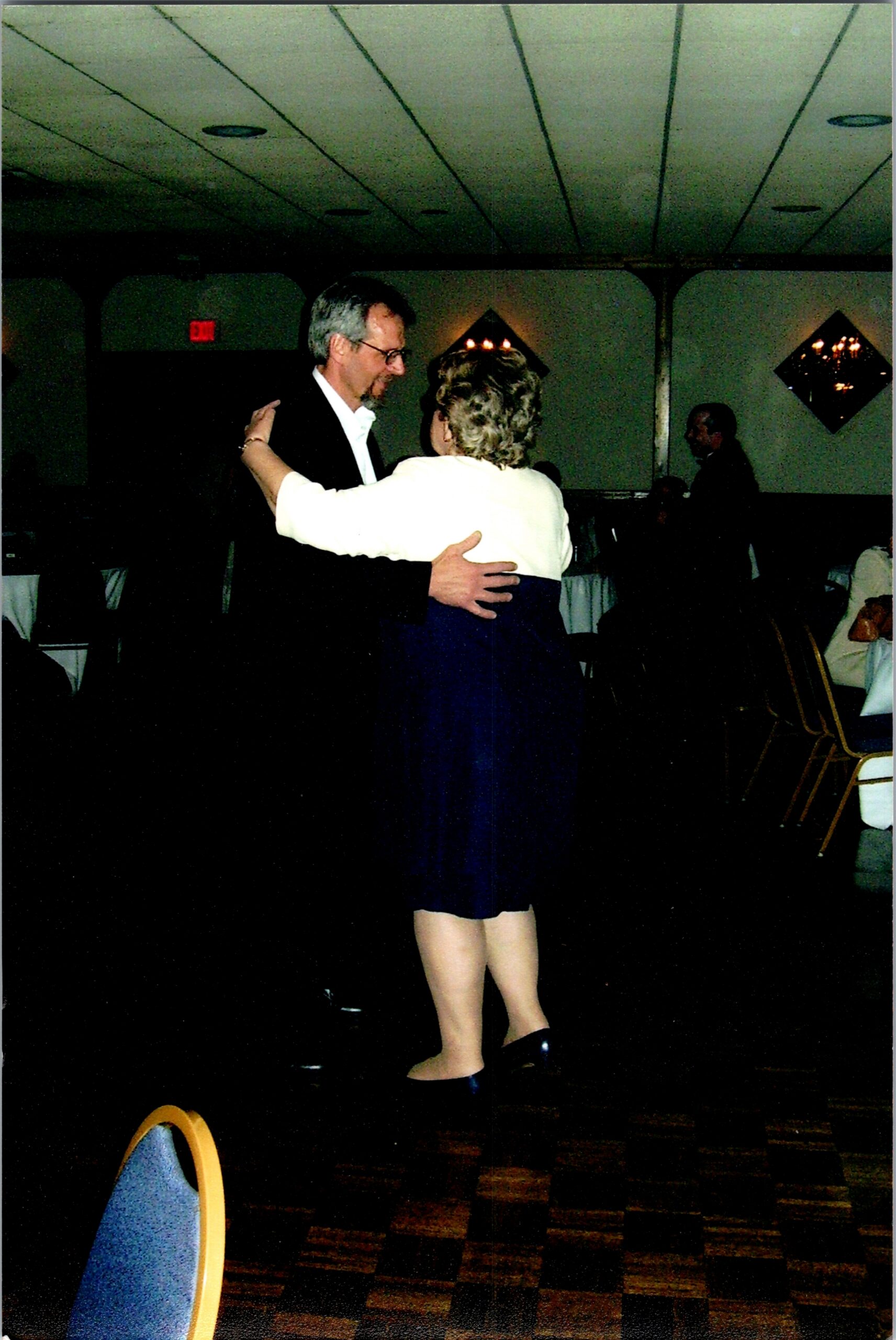 Couple dancing closely at a formal event in a dim banquet hall; man in a dark suit, woman in white top and navy skirt, others seated in background.