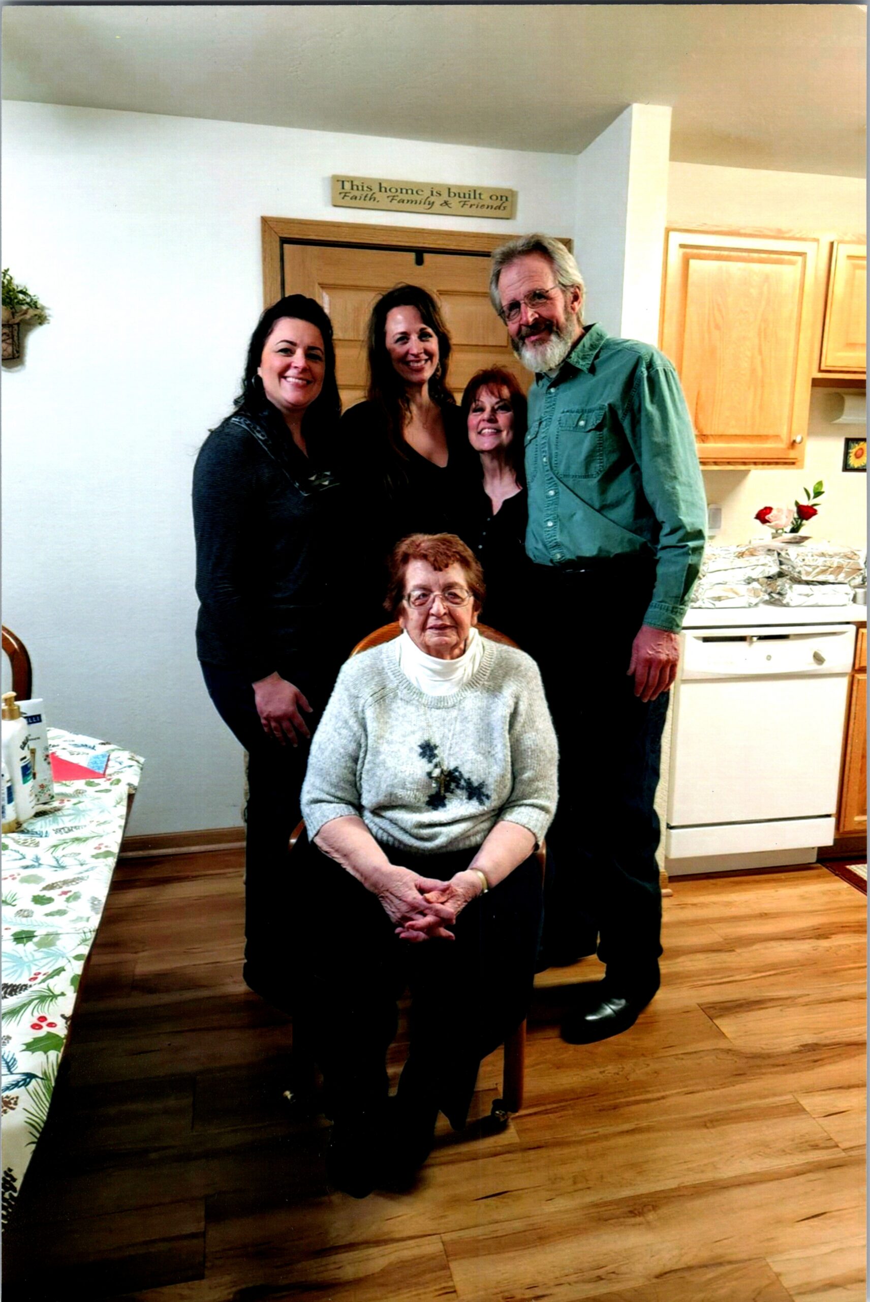 Group of five smiling people posing in a cozy kitchen; an elderly woman sits in front, others stand behind her near wooden cabinets and a stove. A sign reads: 'This home is built on Faith, Family & Friends'