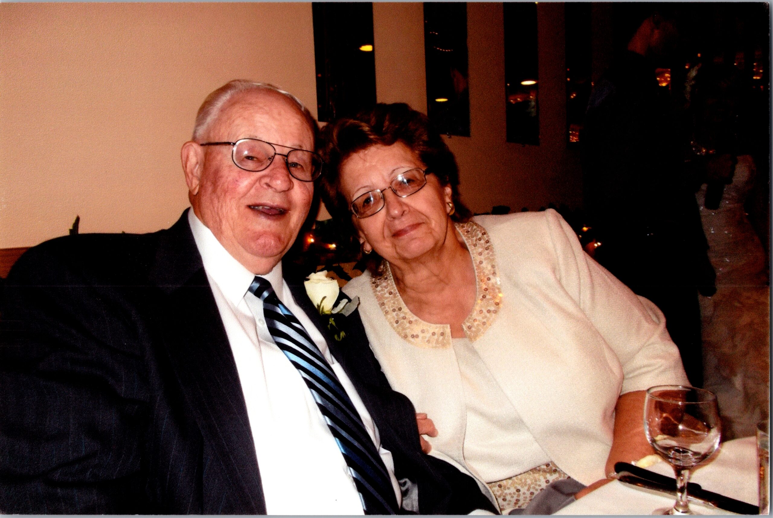 Senior couple posing for a photo at a formal dinner, smiling at the camera; the man wears a boutonniere and a striped tie.