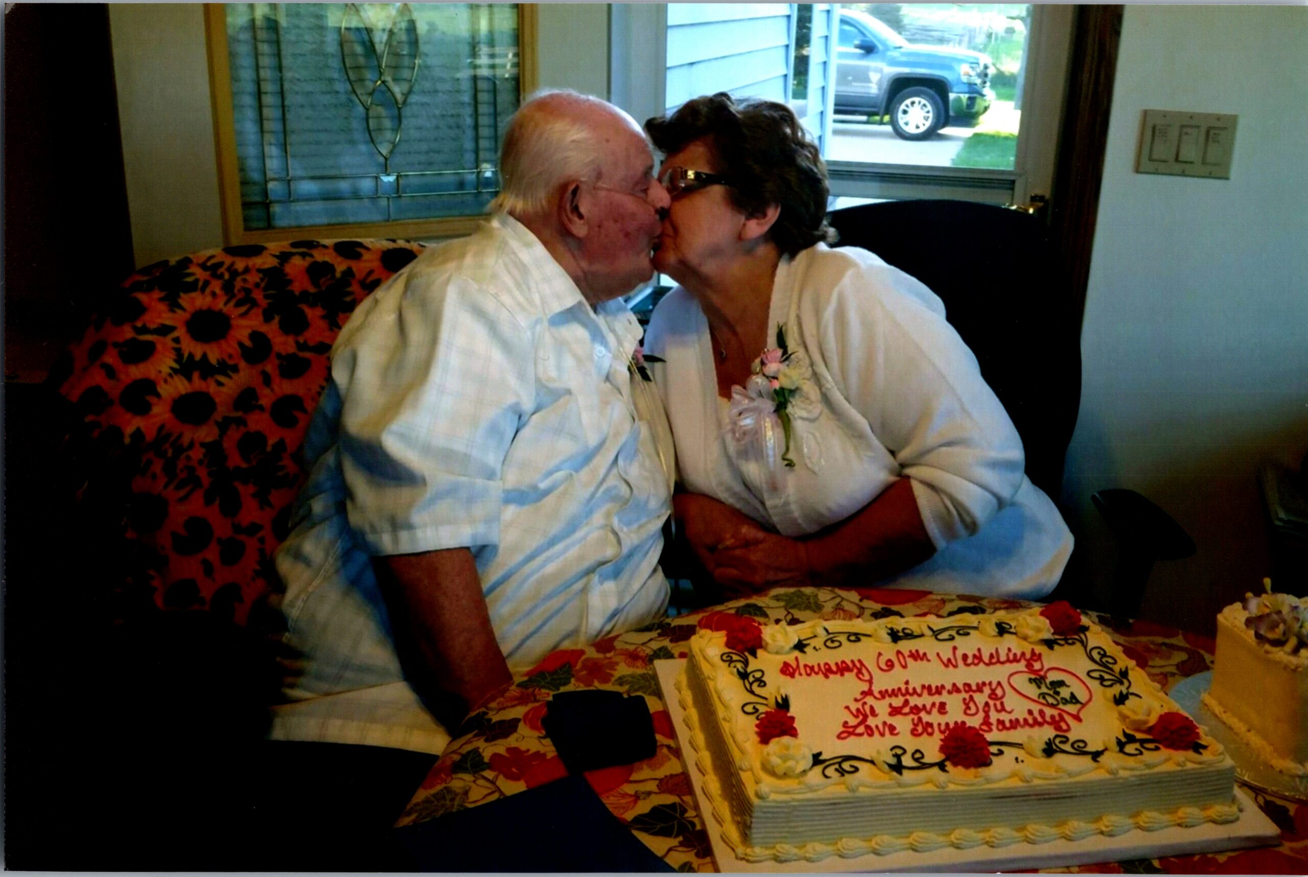Elderly couple kissing beside a decorated anniversary cake on a floral tablecloth indoors, with a window in the background.