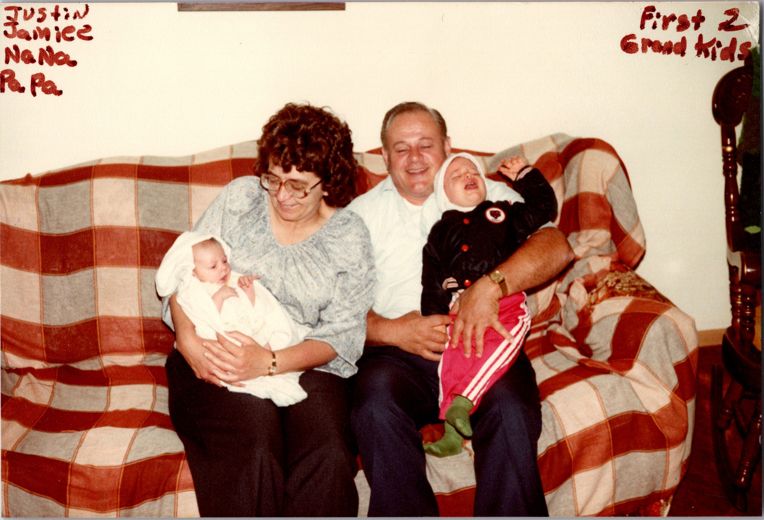 Grandmother with a baby and grandfather with a crying toddler sitting together on a red-and-tan plaid sofa at home.