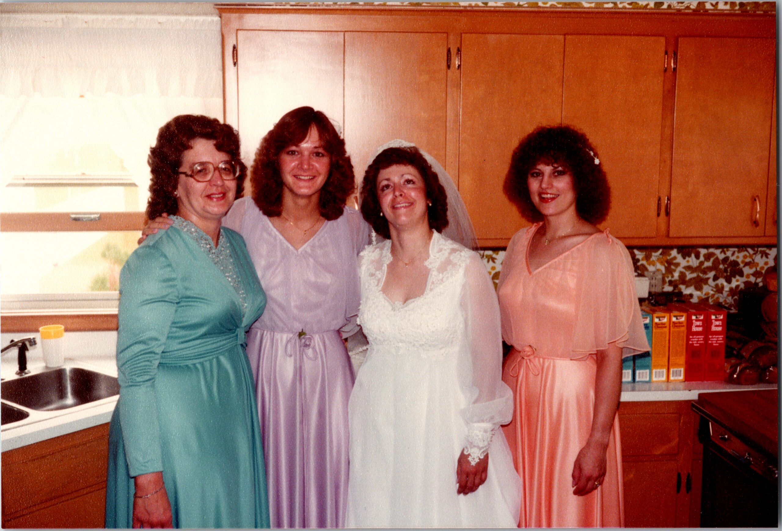 Four women in bridesmaid dresses pose together in a kitchen, with wood cabinets and floral wallpaper in the background.