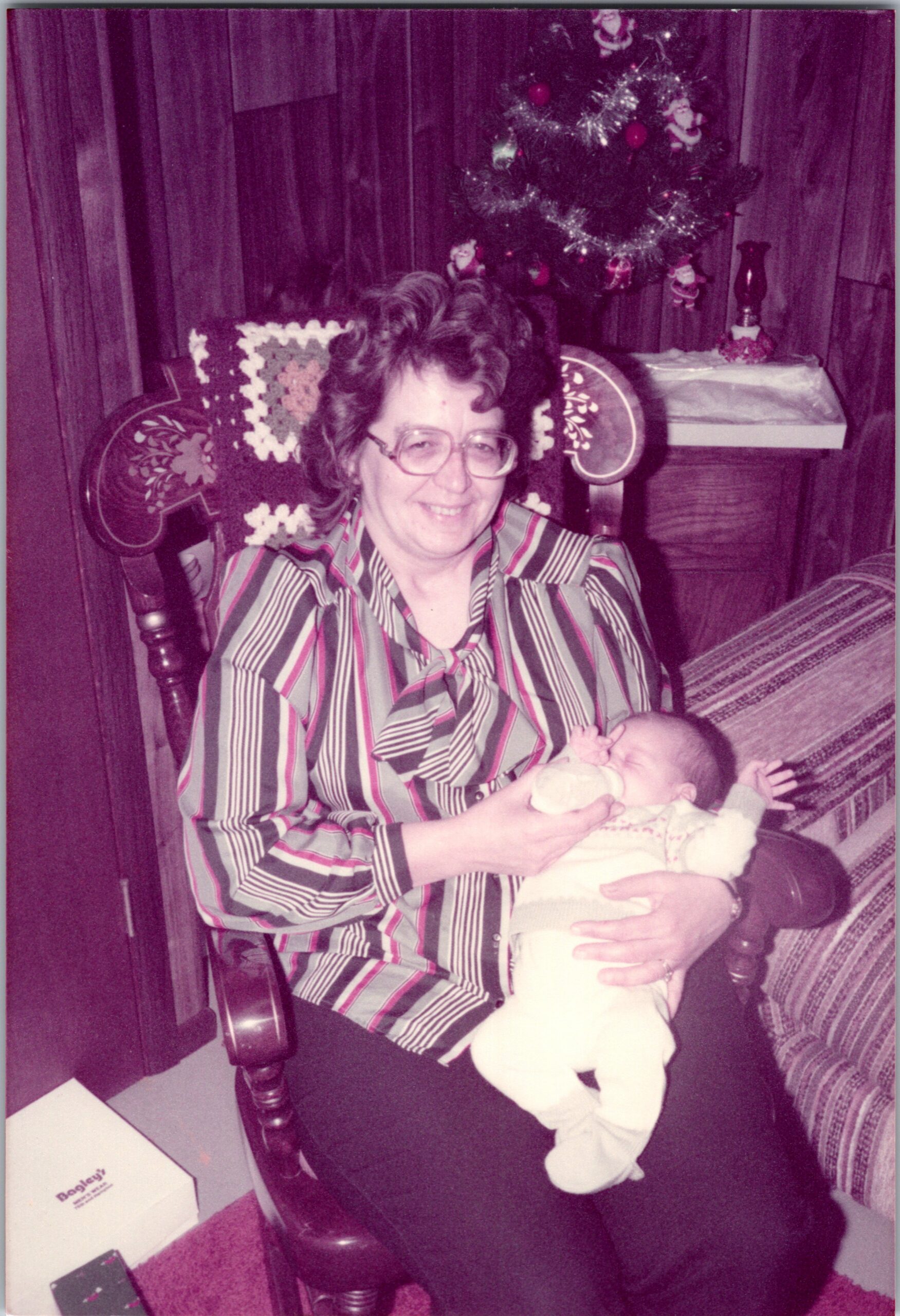 Smiling woman in a striped shirt sits in a wooden chair, feeding a baby with a bottle; a Christmas tree and decorations are in the background.