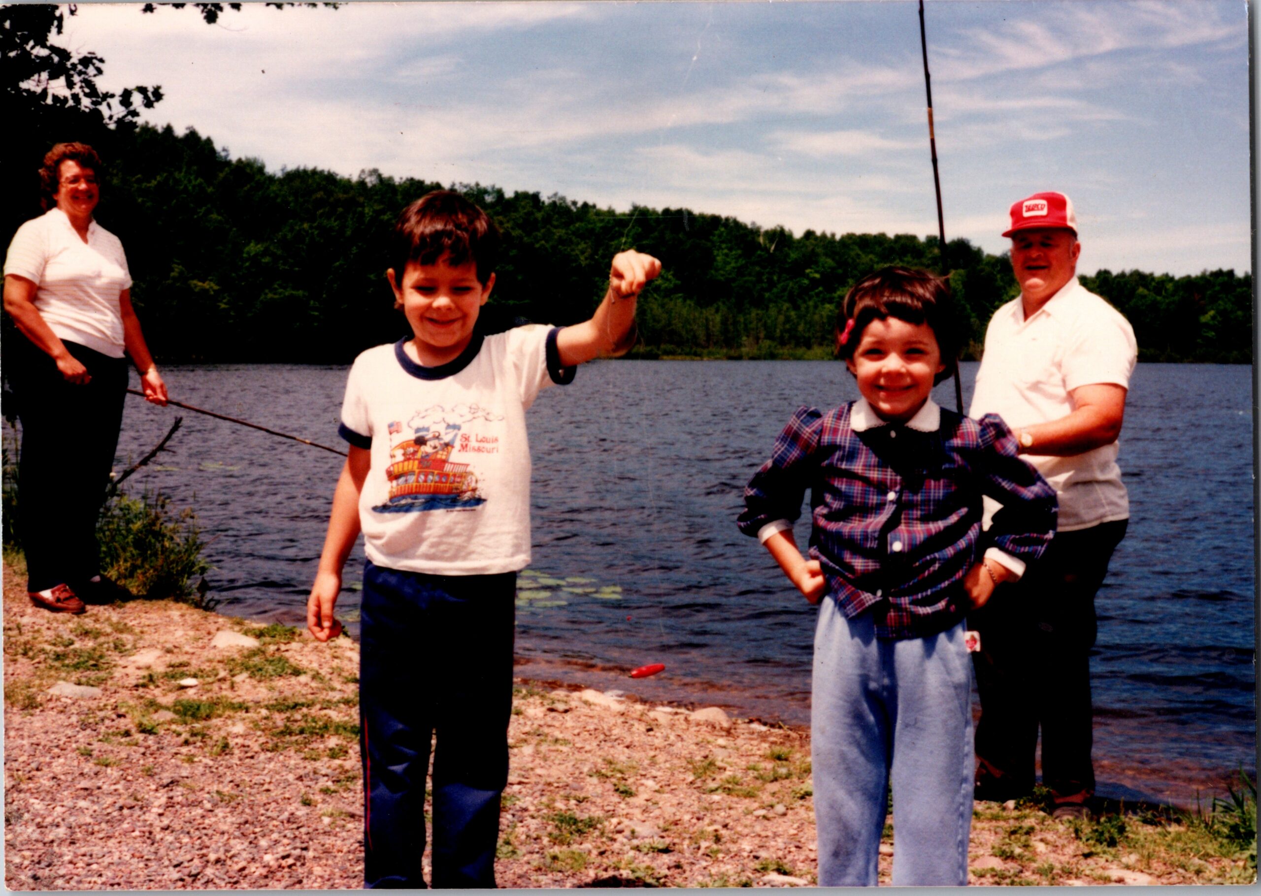 Family by a lake: two children posing at the shore with a man and woman nearby, trees in the background on a sunny day.