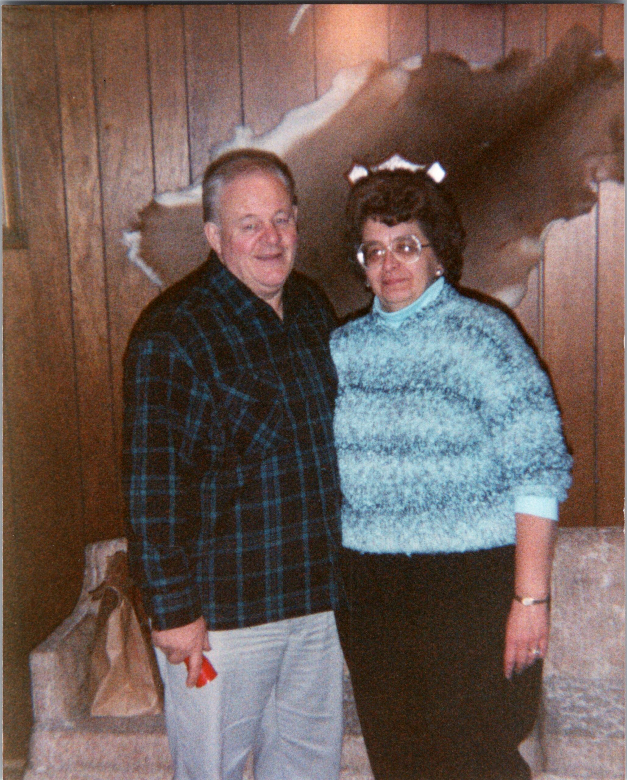 An older man in a dark plaid shirt stands beside an older woman in a light blue knit sweater and glasses, both smiling for the photo indoors with wood-paneled walls behind them.
