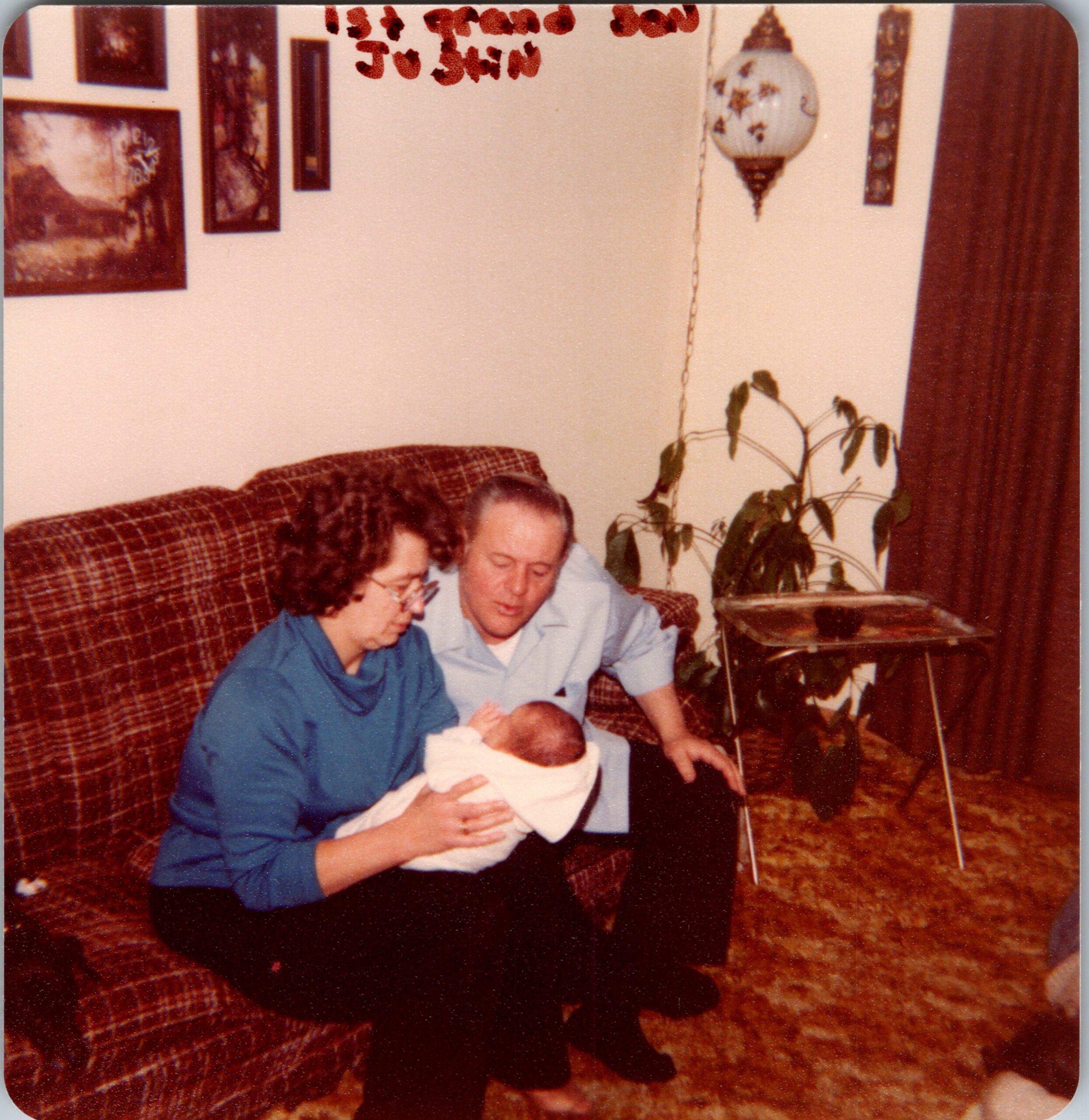 Older couple seated on a plaid sofa, gazing at a newborn baby in a cozy living room. A plant and wall decor are visible in the background.